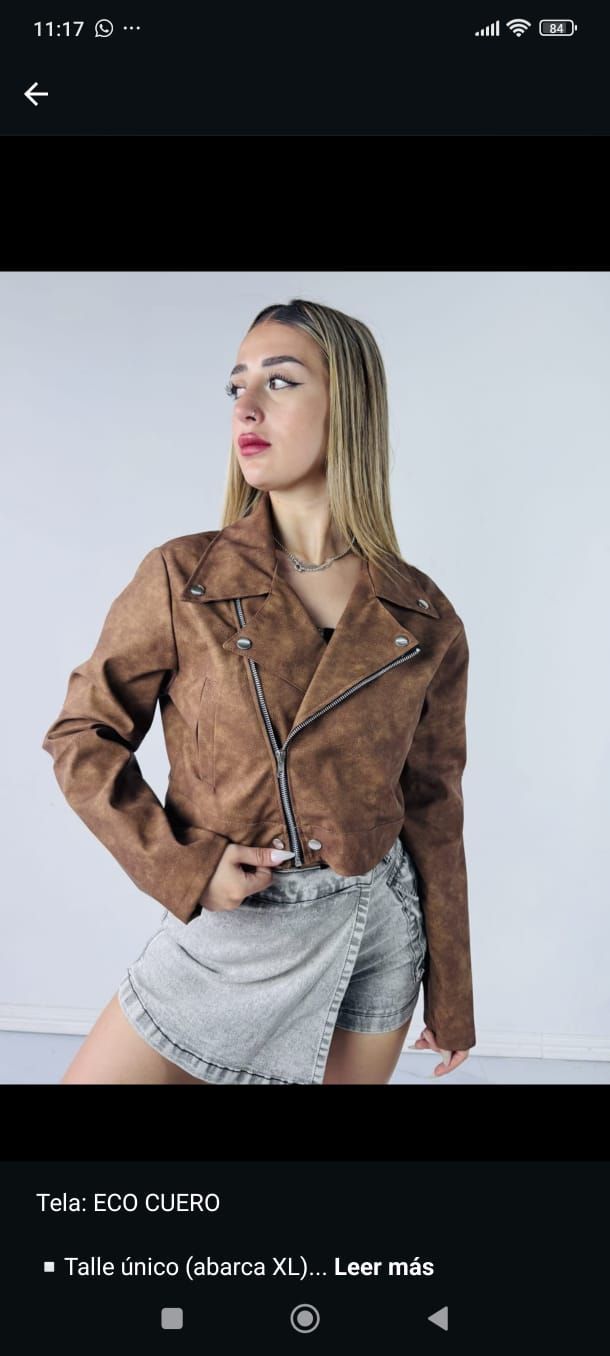 Mujer rubia con chaqueta marrón y falda gris posando frente a un fondo gris claro.