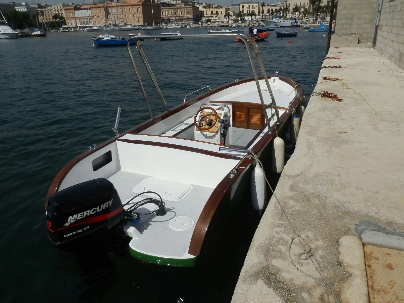 Piccola imbarcazione a motore bianca ormeggiata accanto a una banchina di pietra in un porto con acque calme ed edifici sullo sfondo.