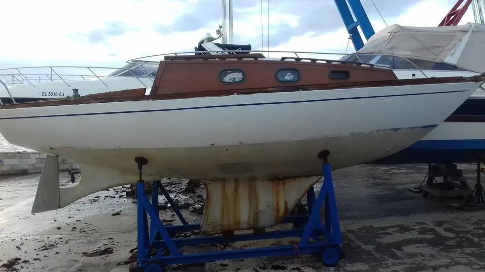 Barca a vela su un cavalletto blu in un cantiere navale, scafo bianco con cabina marrone e alberi sullo sfondo.