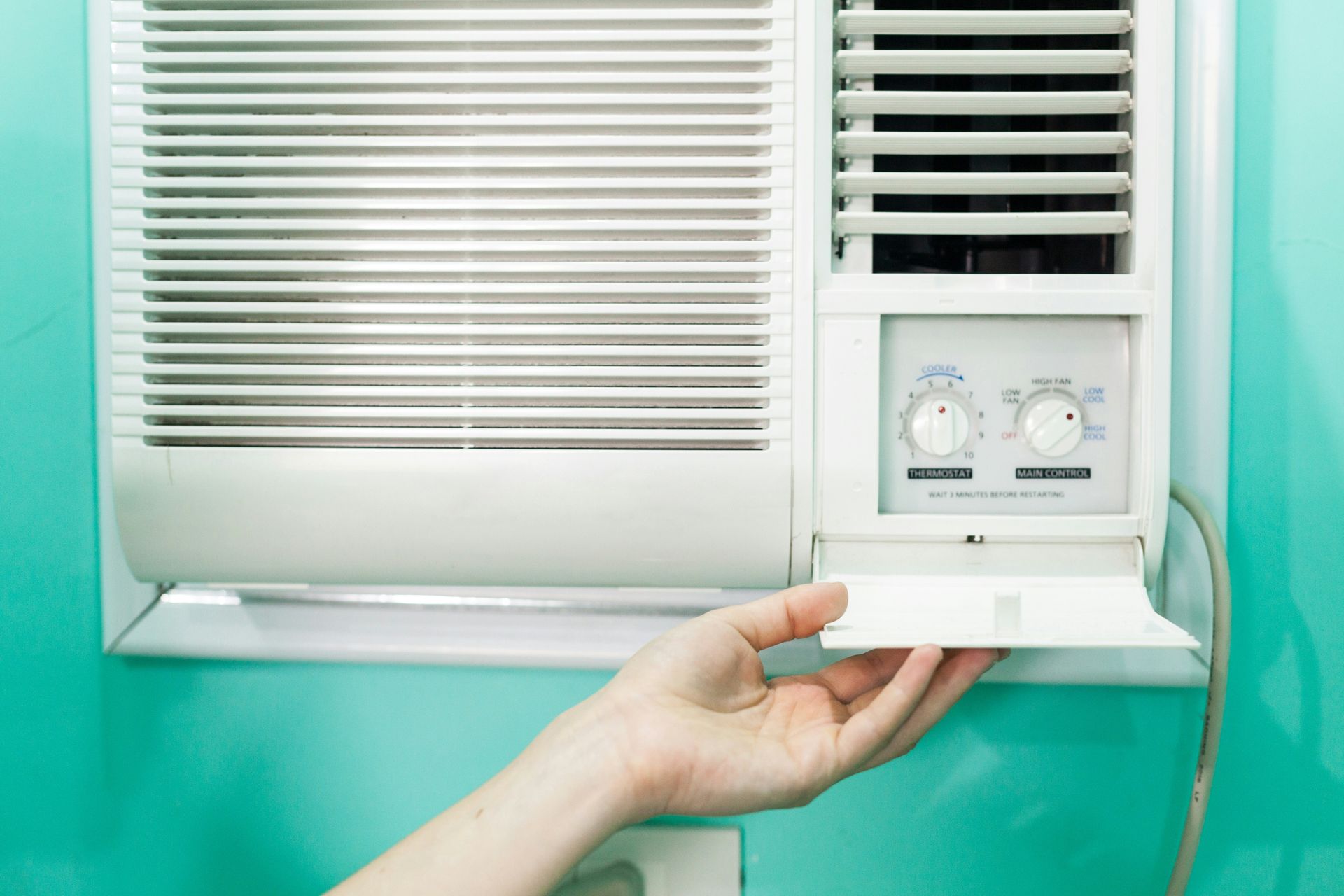 Hand adjusting a window air conditioner’s control panel beside closed blinds