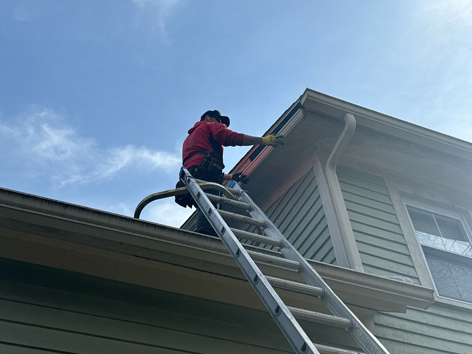 A person in a red jacket standing on a ladder, cleaning the gutters of a light green house against a blue sky.