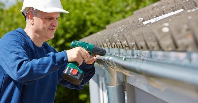 A person wearing a white hard hat uses a power drill to install a metal gutter along the edge of a shingled roof.