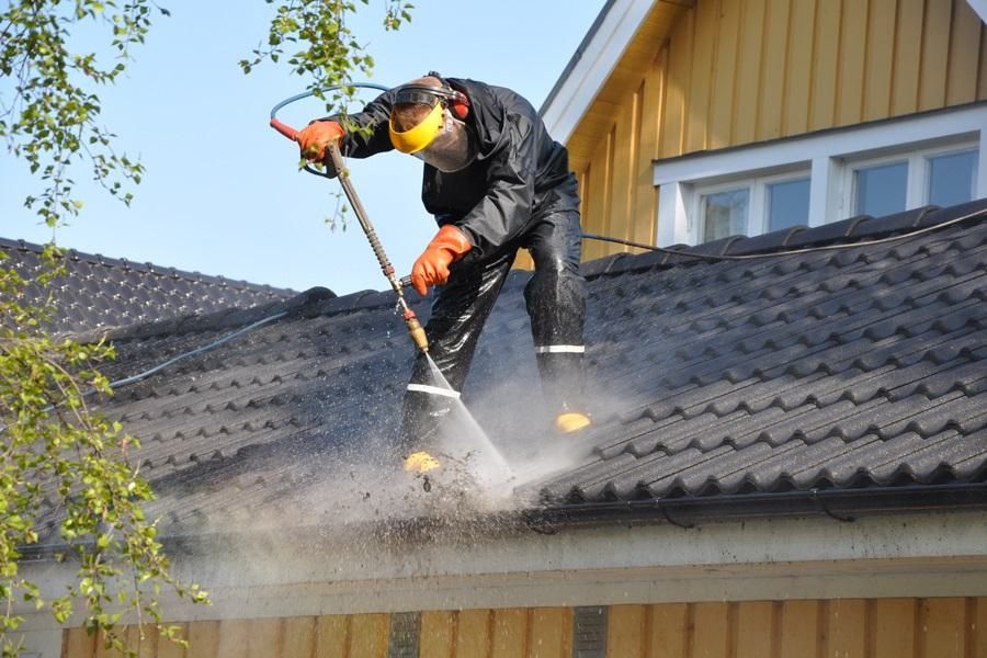 A worker in protective gear power washes a dark, tiled roof on a yellow house.