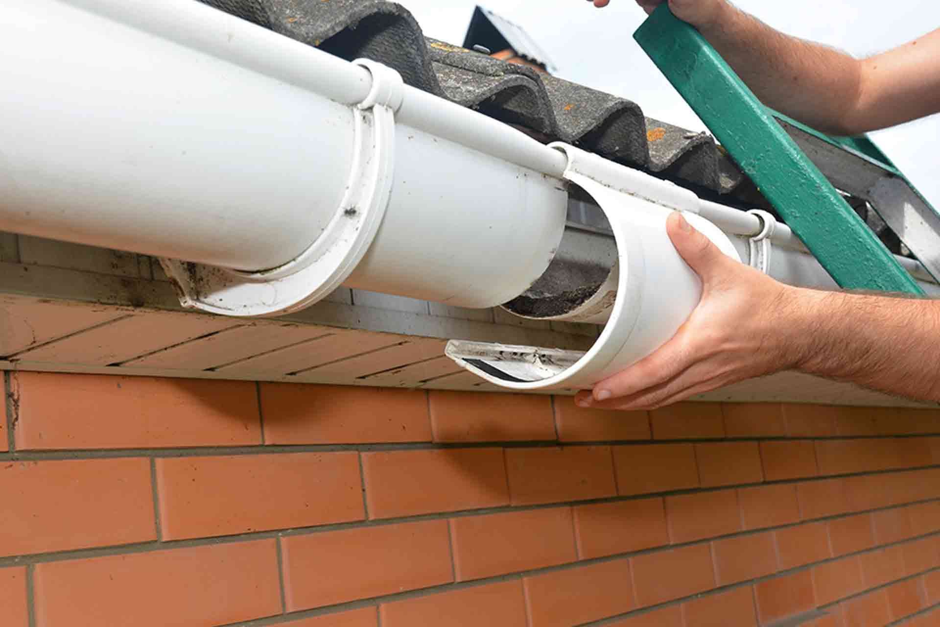 A person standing on a ladder and connecting white plastic sections of a rooftop gutter system on a brick building.
