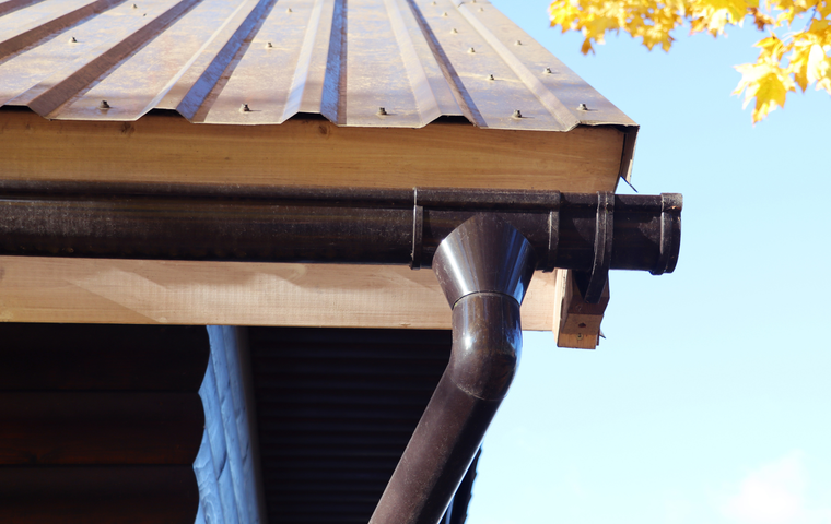 A brown metal roof with a matching gutter system and downspout attached to a wooden fascia board under a clear blue sky.
