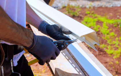 A worker wearing gloves uses tin snips to cut a piece of white vinyl siding outdoors.