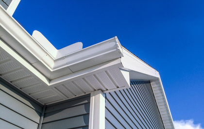 White gutters and ventilated soffits on the exterior corner of a house with grey siding against a clear blue sky.