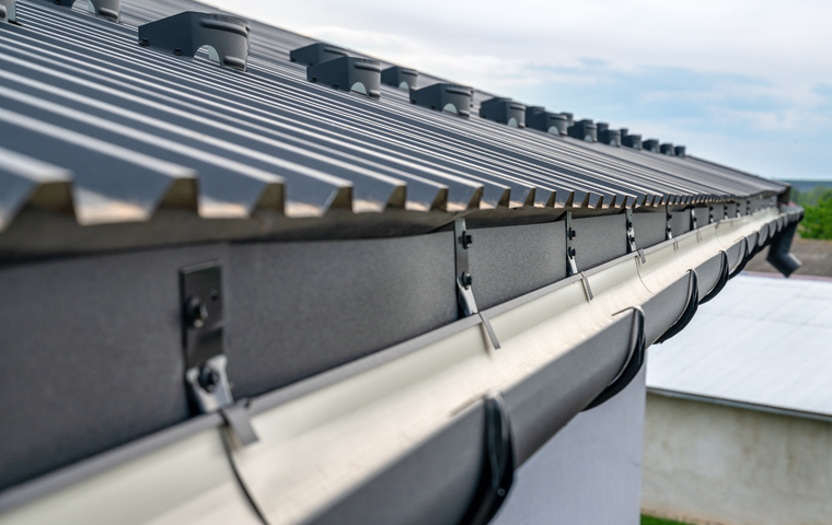Close-up of a metal roof edge with a white gutter system mounted to the side of a building under a cloudy sky.