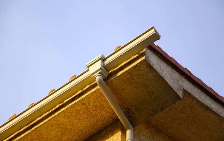 A tan rain gutter attached to the edge of a roof against a clear blue sky.
