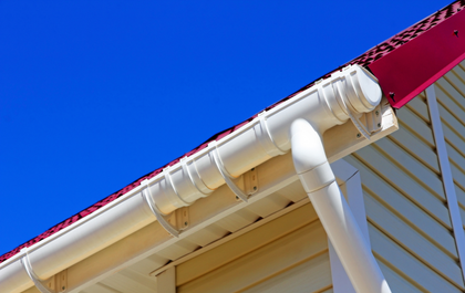 White plastic rain gutter and downspout attached to the side of a building with red roofing under a clear blue sky.