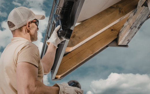 A worker wearing a cap and gloves attaches a black gutter to the wooden eaves of a building against a cloudy sky.