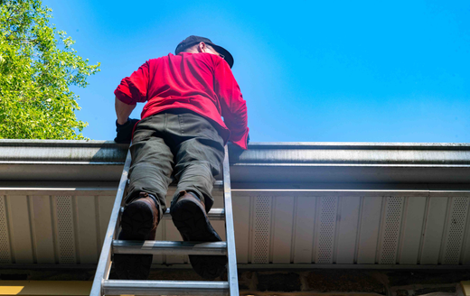 A person in a red shirt and dark pants stands on an extension ladder while cleaning a home's gutters against a blue sky.