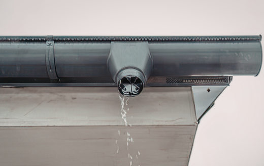 A grey metal roof gutter section with a downspout outlet dripping water over a house fascia board.