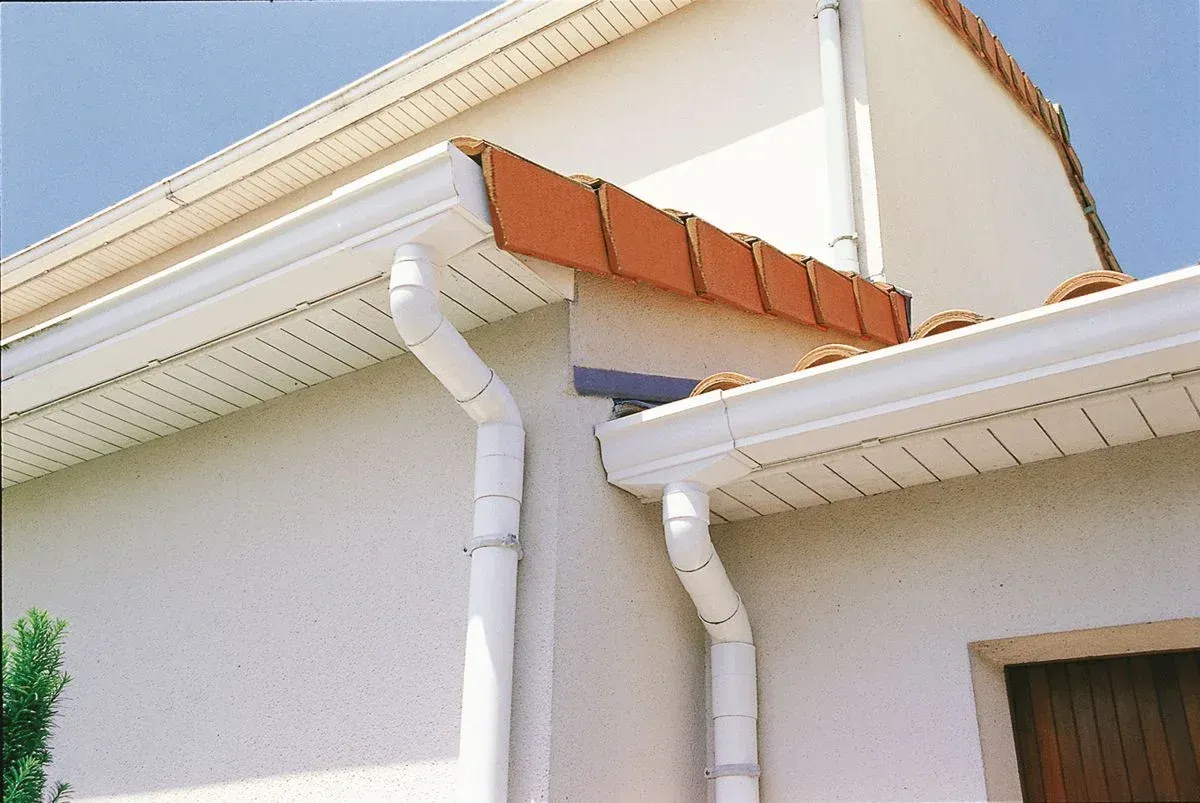 White roof gutters and downspouts installed on the exterior corner of a beige house with a tiled roof.