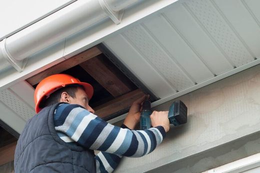 A person wearing an orange hard hat and vest uses a power drill to install soffit panels under a building's roof overhang.