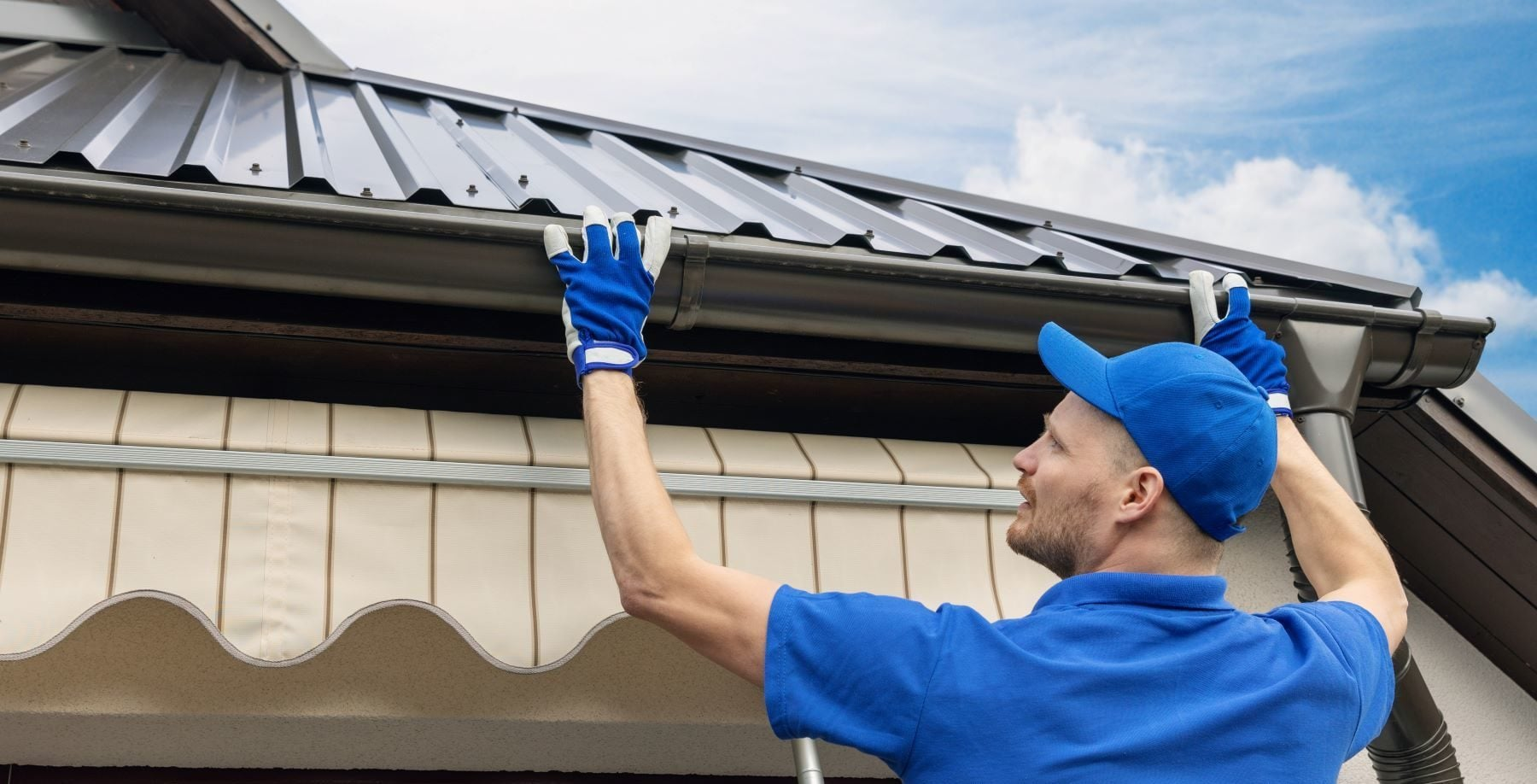 A person in a blue uniform and gloves cleans a house gutter along a corrugated metal roof.