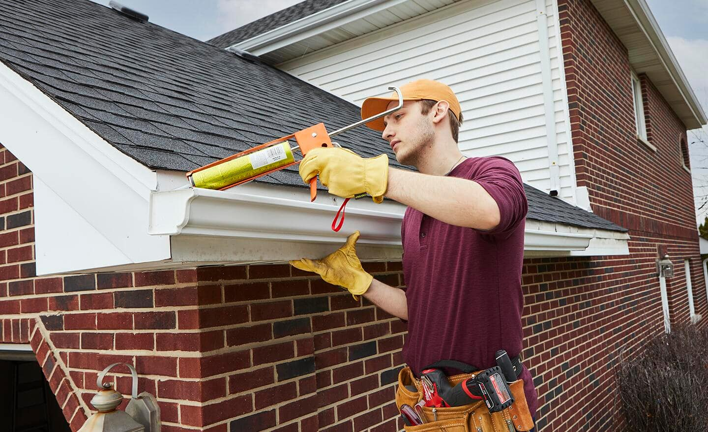 A person in a red shirt and yellow gloves uses a caulking gun to seal the edge of a white gutter on a brick house.