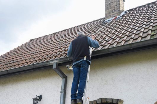 A person climbs an extension ladder leaned against the side of a house with a tiled roof and a chimney.