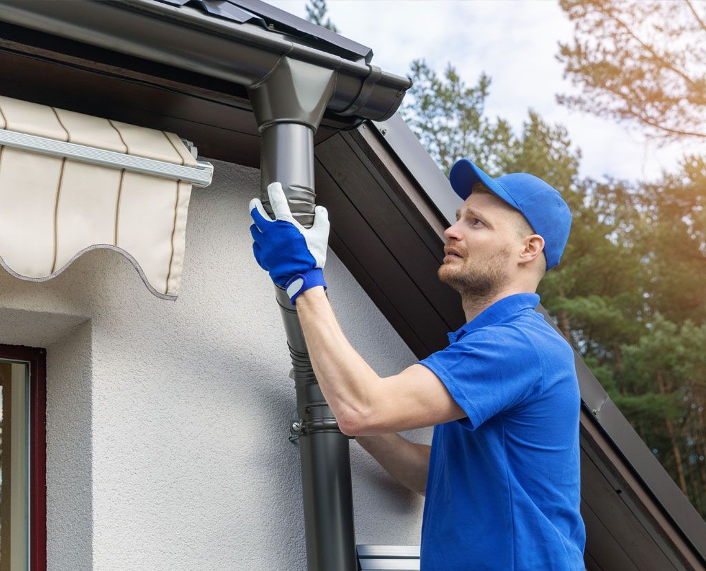 A worker in a blue uniform and gloves attaches a dark gutter downspout to the side of a house.