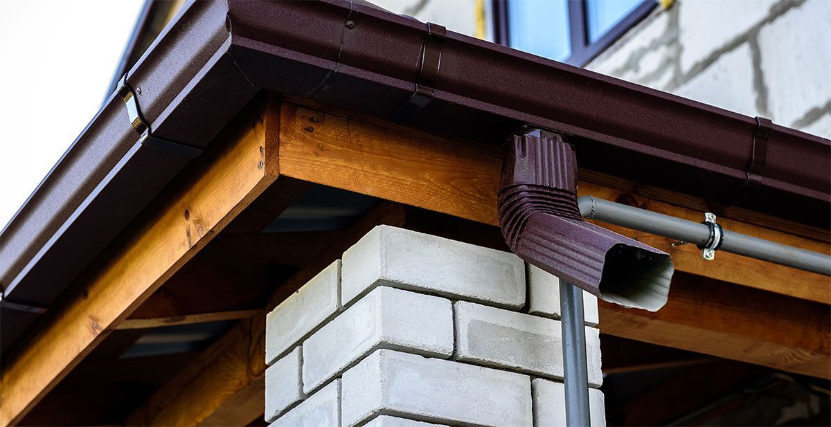 A brown metal rain gutter and downspout attached to a wooden roof eave next to a white brick pillar.
