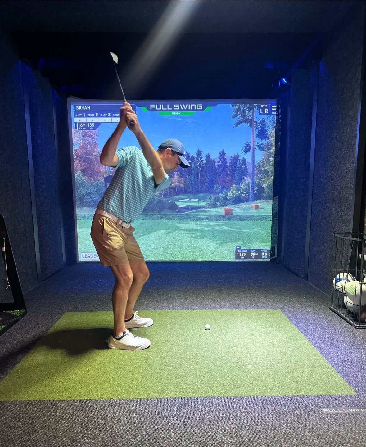 Man swings golf club in indoor golf simulator; green mat, screen displays golf course.