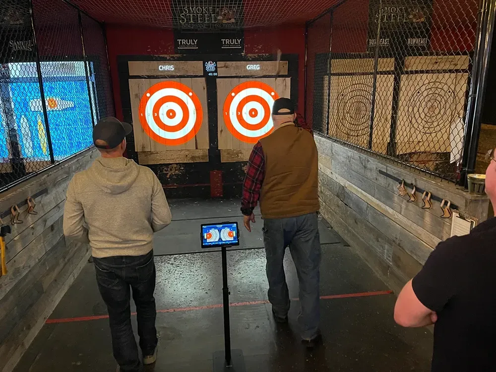 Men throwing axes at targets in an axe-throwing venue. Two targets with orange rings, and a digital scoreboard.