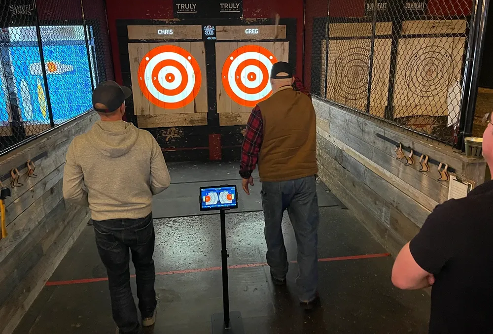 Men throwing axes at targets in an axe-throwing venue. Two targets with orange rings, and a digital scoreboard.