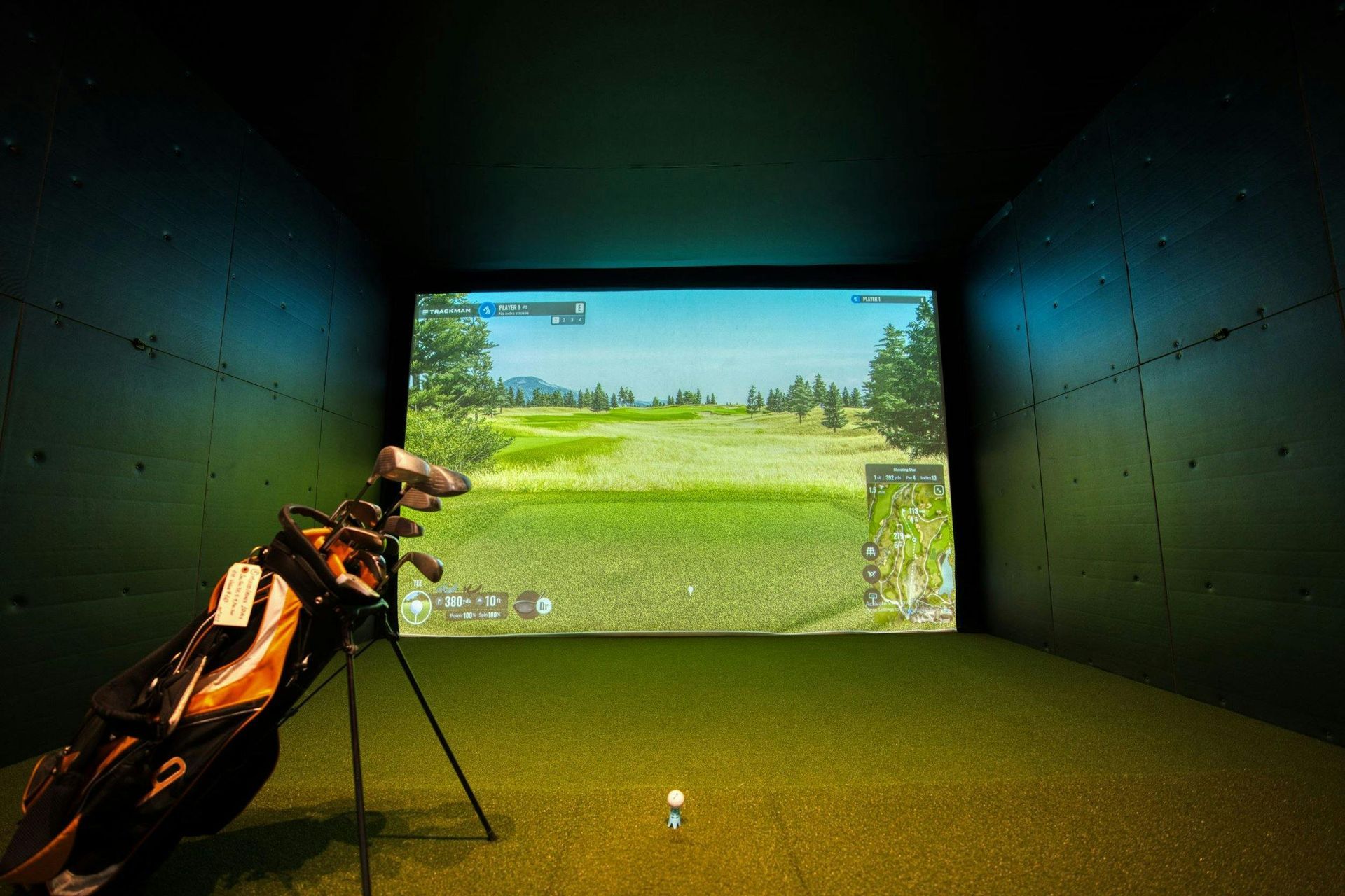 Golf simulator room with a golf bag, a ball, and a screen displaying a golf course.