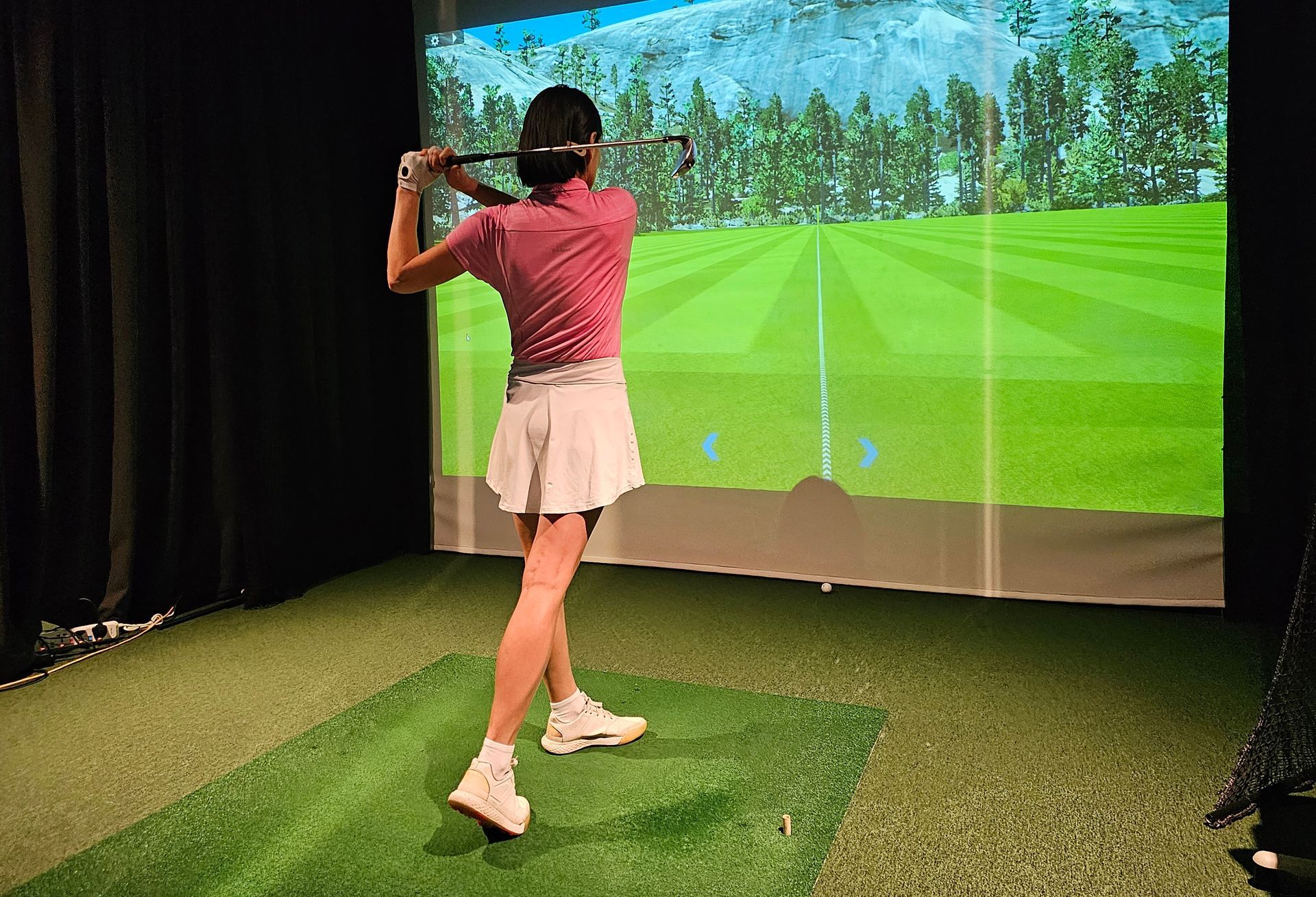 Woman in pink shirt and white skirt swings golf club in indoor simulator. Green mat, screen background.