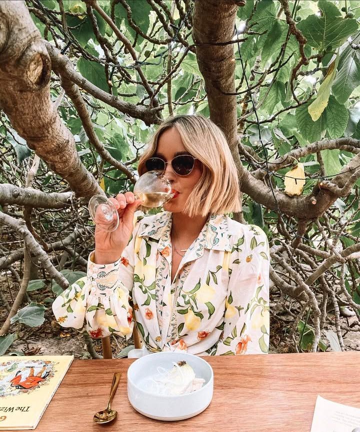 Woman drinks wine outdoors, wearing sunglasses and floral top, seated at a table with a small dessert