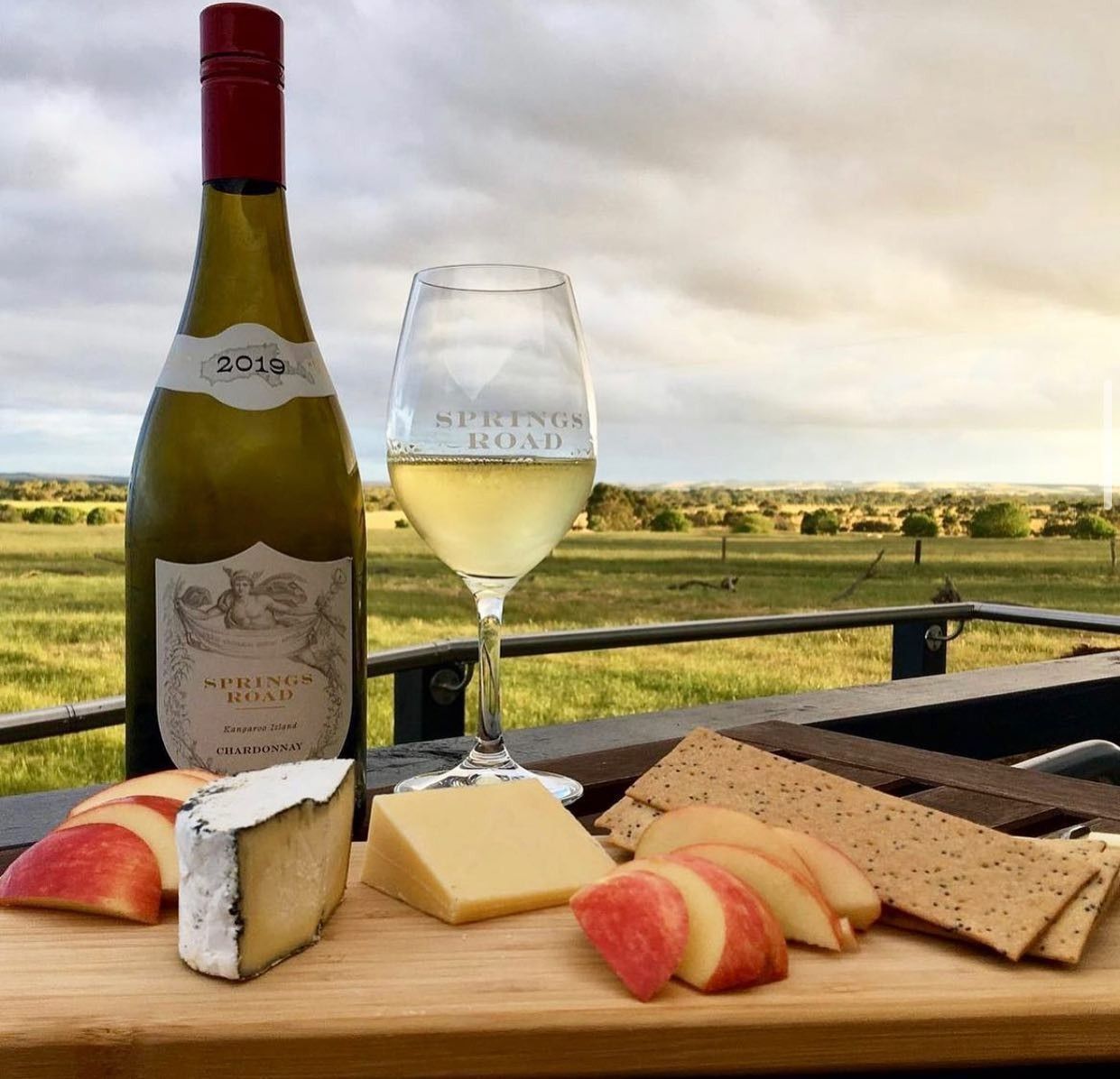 A bottle of white wine, a glass of wine, cheese, crackers, and apples on a wooden board outdoors.