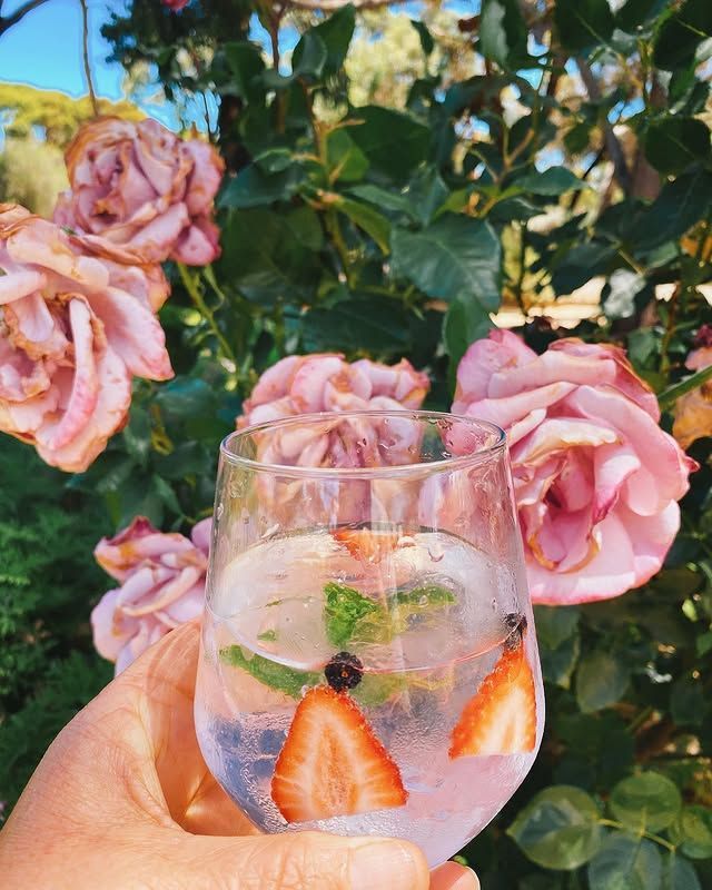 Hand holding a glass of cocktail with strawberries and mint, roses in the background.