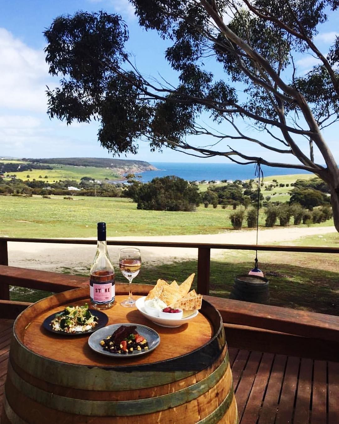 A wine barrel table on a deck with food and wine overlooking a scenic green landscape and ocean.
