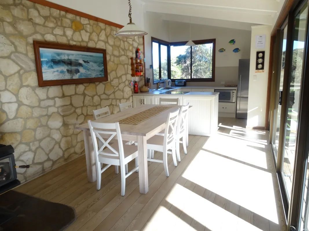 Bright dining area with white table and chairs, stone wall, and open window overlooking greenery