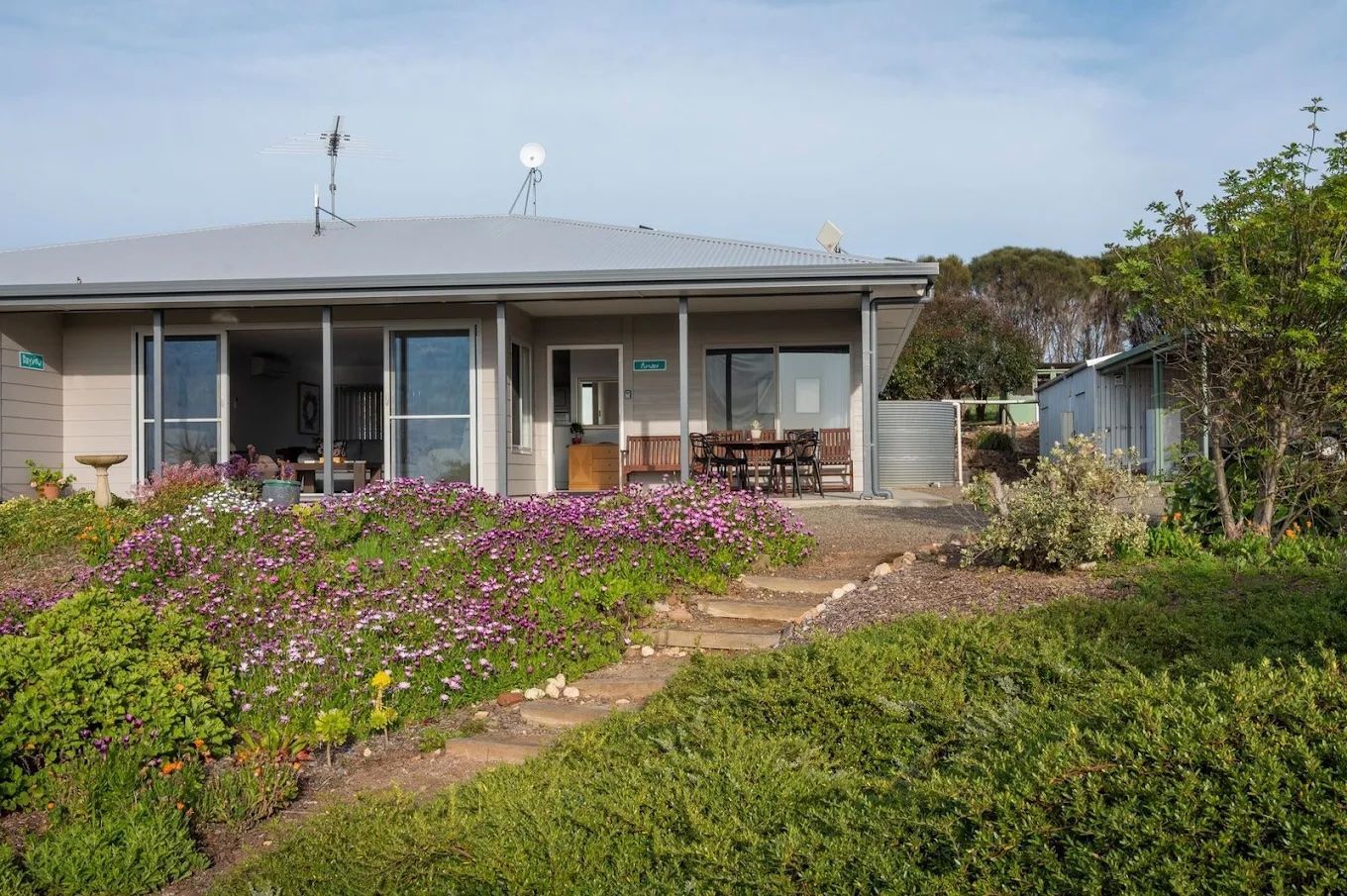Single-story house with a front porch, steps, and a flower-filled garden under a gray roof.