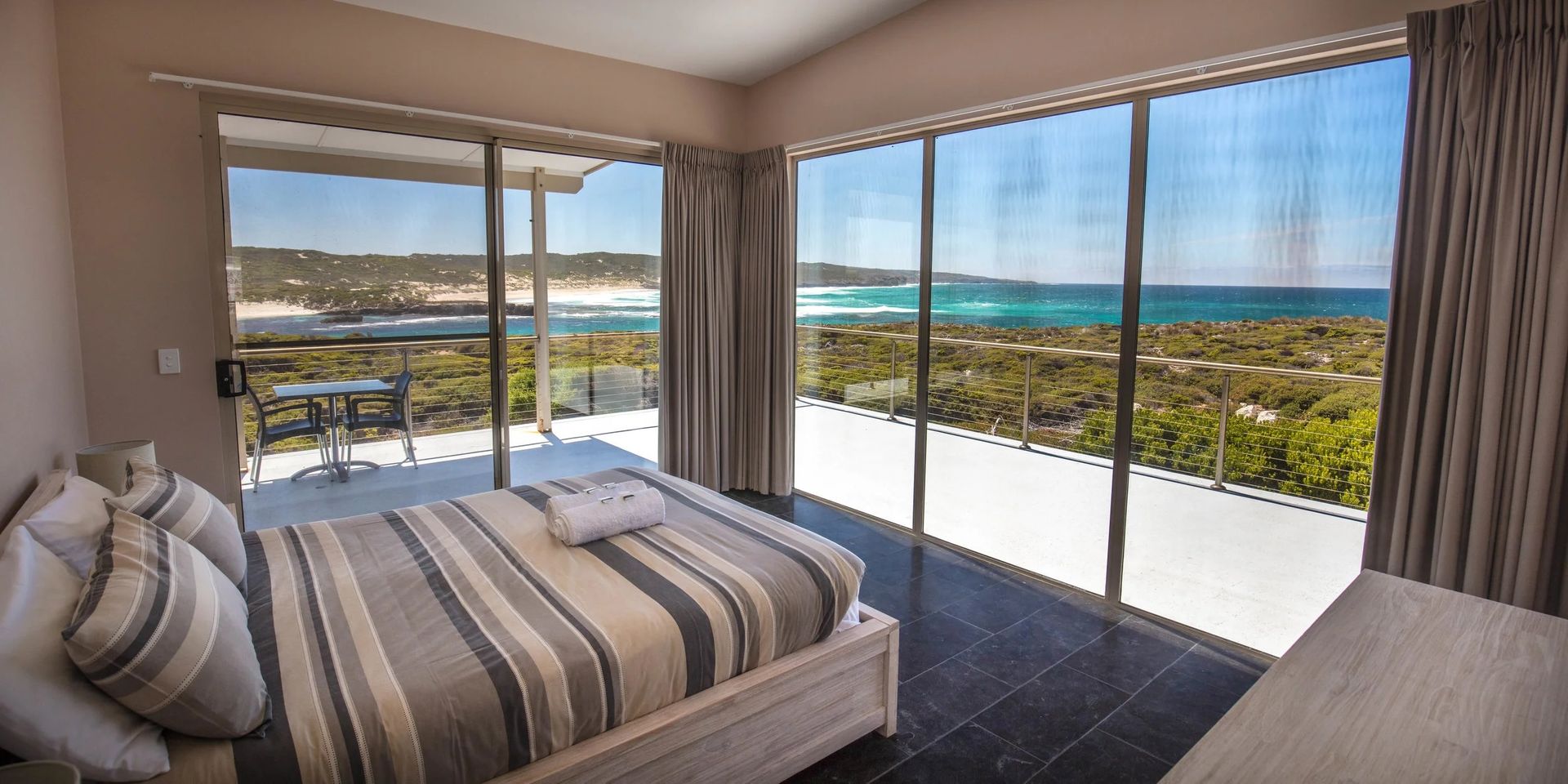 Bright bedroom with striped bed, floor-to-ceiling windows, and ocean view from a balcony