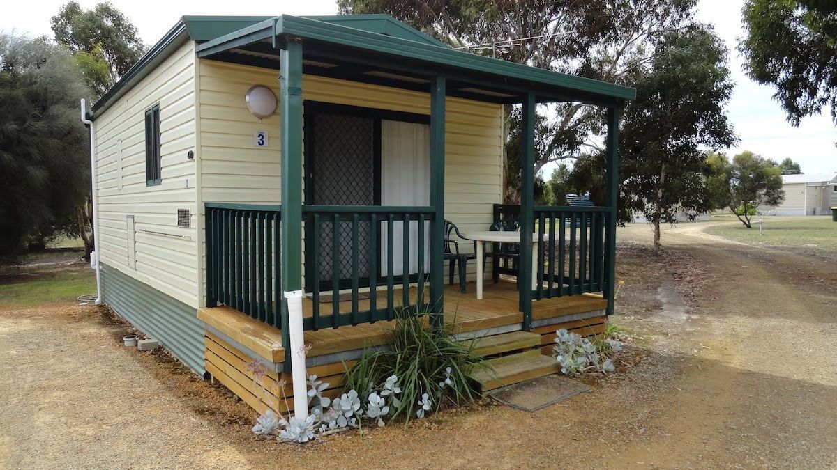 Small beige cabin with green porch railings and steps, set on a gravel lot with trees nearby.