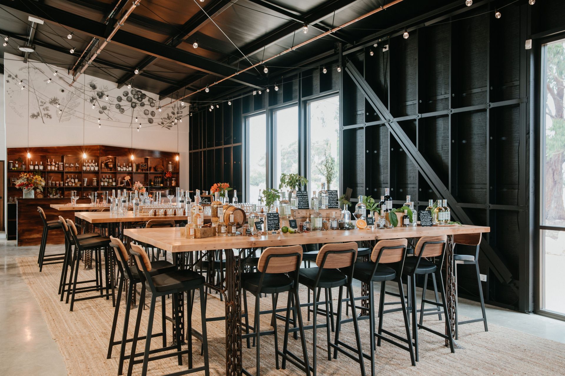 Interior of a tasting room with long tables, bar stools, and bottles, with windows and dark walls.