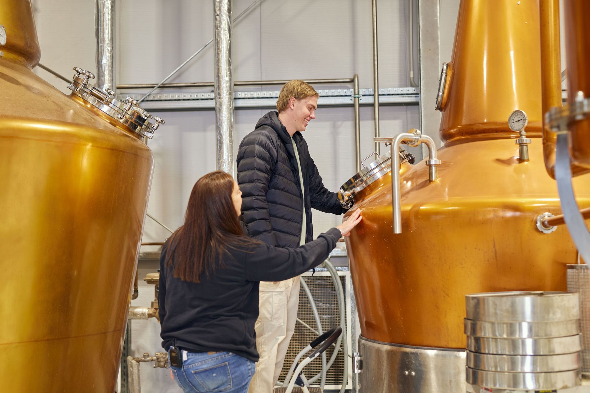 Two people inspecting copper distilling equipment in a distillery.
