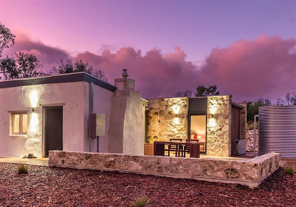 Modern stone house at dusk with warm lights, a patio table, and pink clouds in the sky.