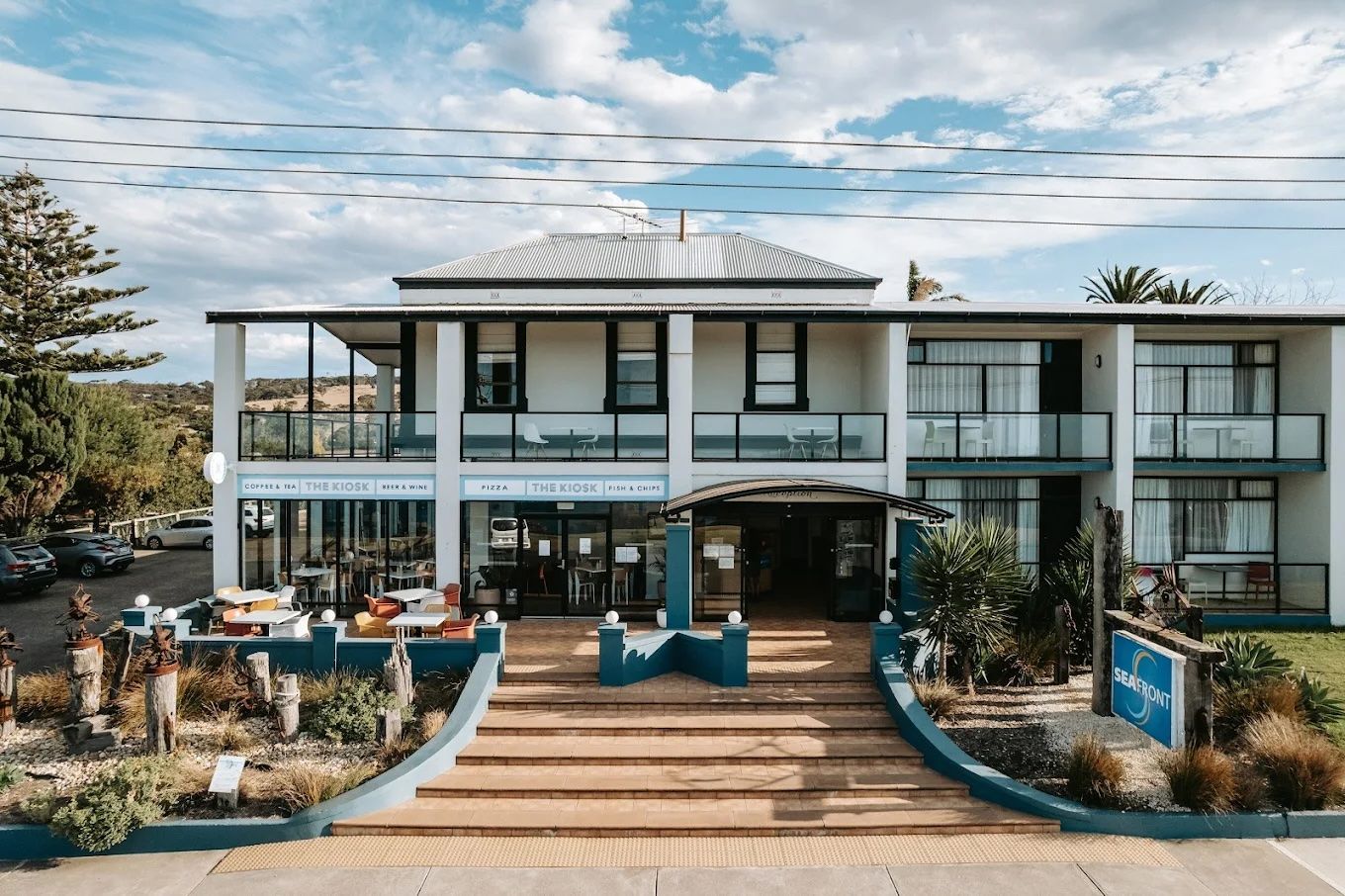 Modern two-story beachfront hotel with a terrace, steps, and outdoor seating under a bright sky