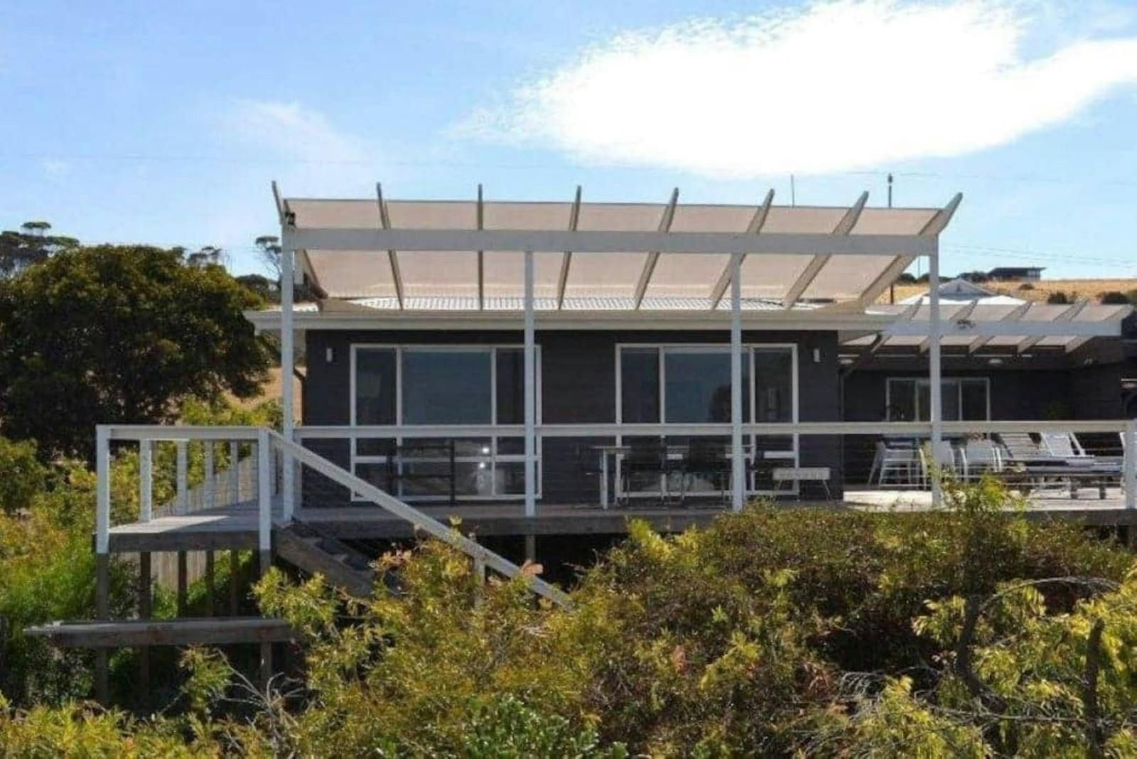 Modern house with large windows and a white roof on a hillside, surrounded by trees under a blue sky