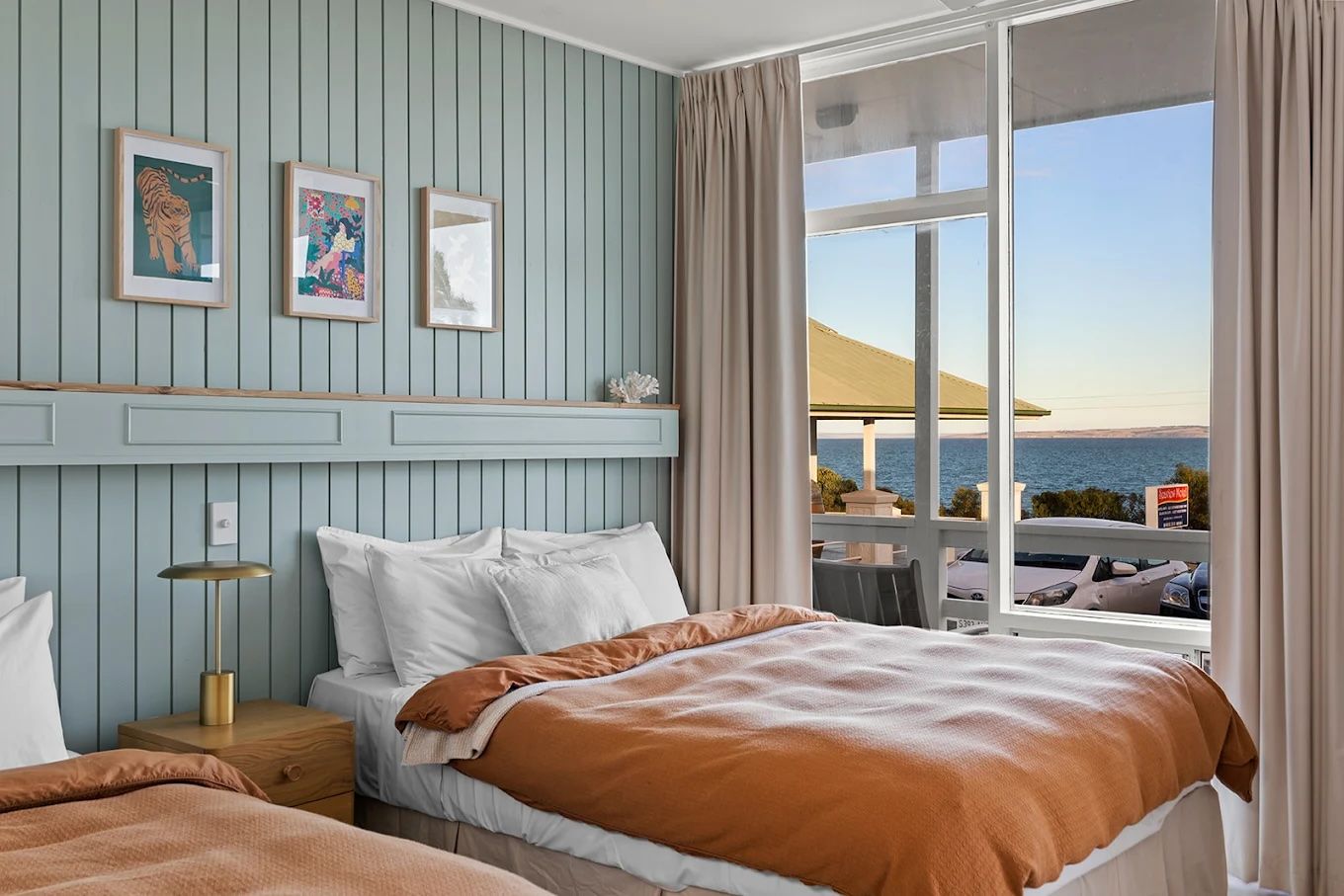 Cozy bedroom with pastel decor, rumpled bed, and balcony view of the sea at sunset