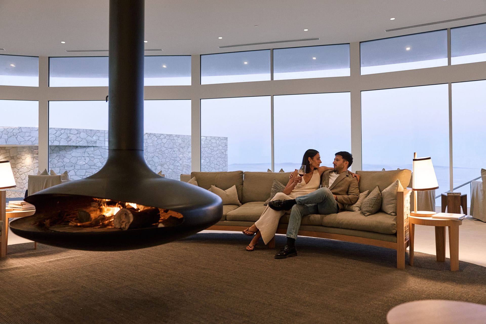 Two people relaxing on a sofa beside a modern fireplace in a bright living room with large windows.
