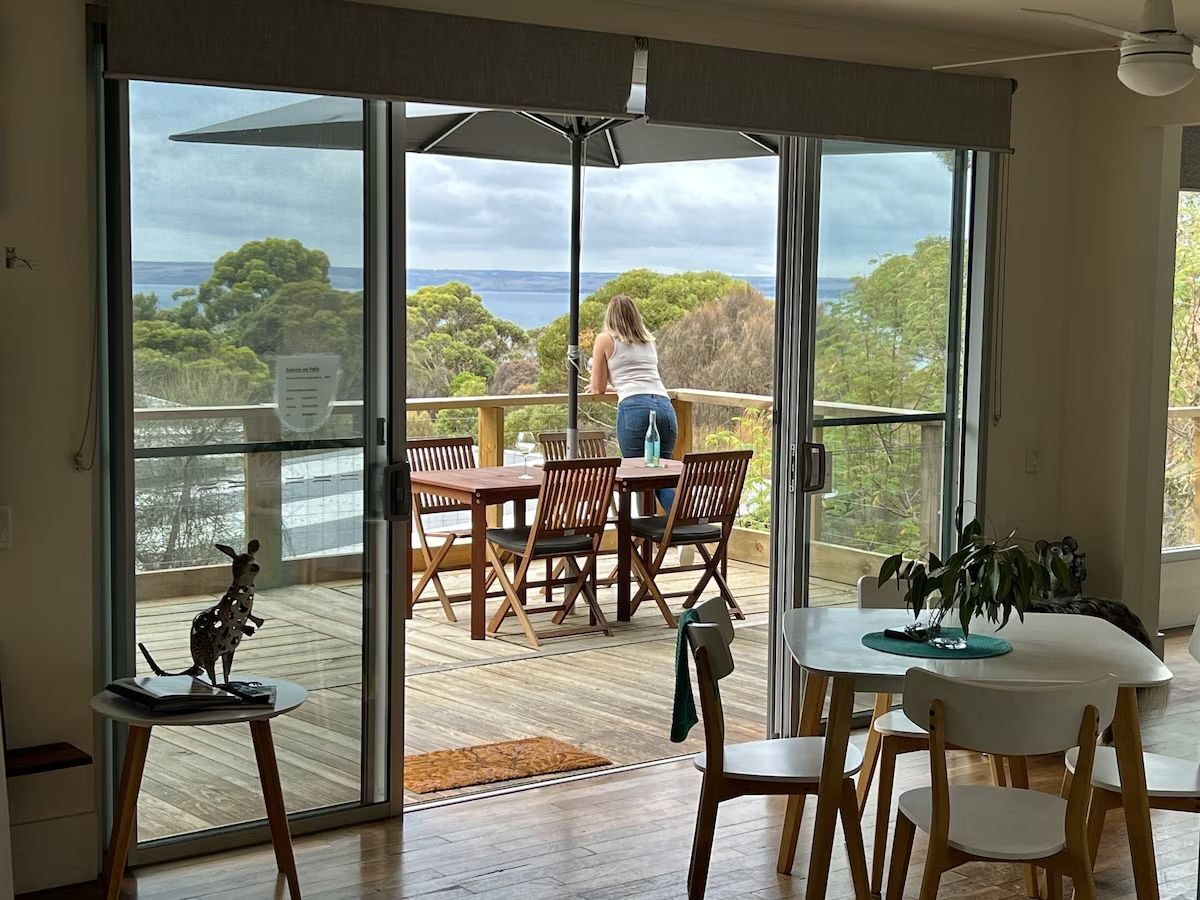 Woman standing on a deck overlooking green trees and water, viewed through open sliding doors.