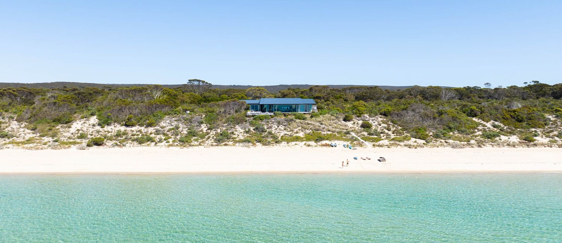Turquoise ocean, sandy beach, and rocky green dunes with a small blue-roofed building on the horizon