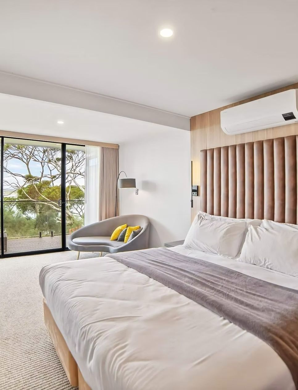 Bright modern bedroom with a large bed, gray chair, wood accents, and a sliding door to greenery outside
