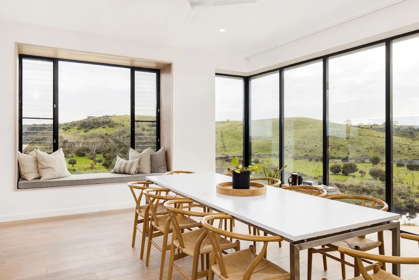 Bright dining room with a long table, wooden chairs, and large windows overlooking green hills