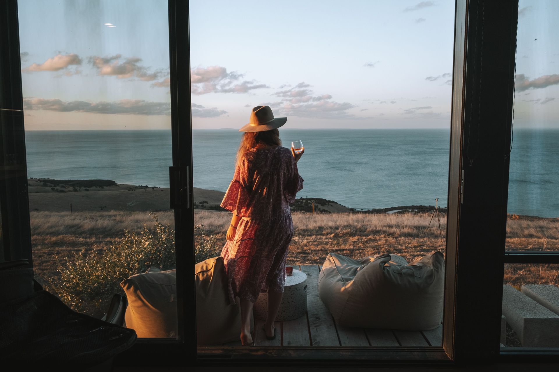 Person in a hat standing on a seaside deck at sunset, looking out over the ocean.