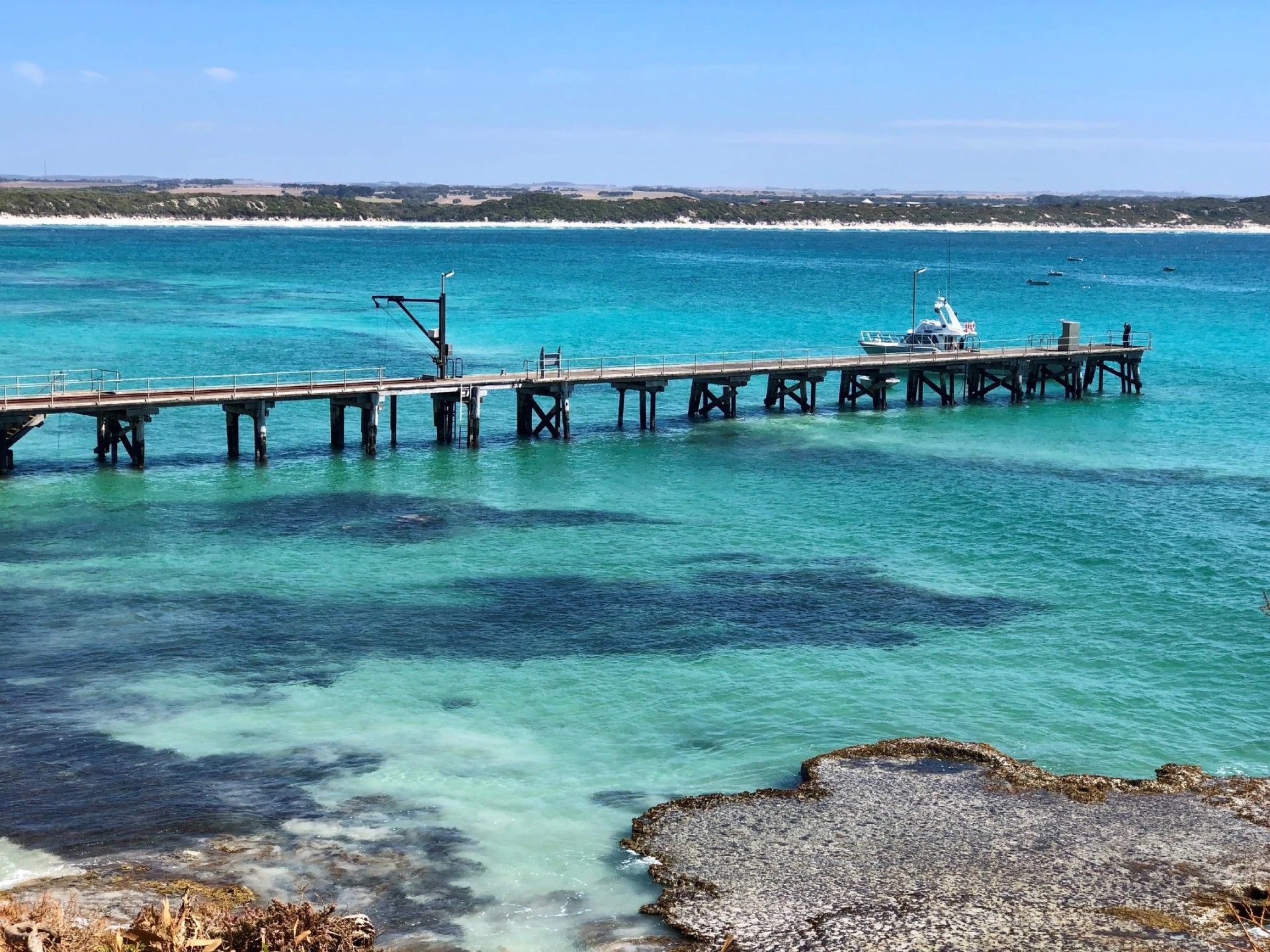 Wooden pier extending into turquoise water, with a boat at the end, and a shoreline in the distance.